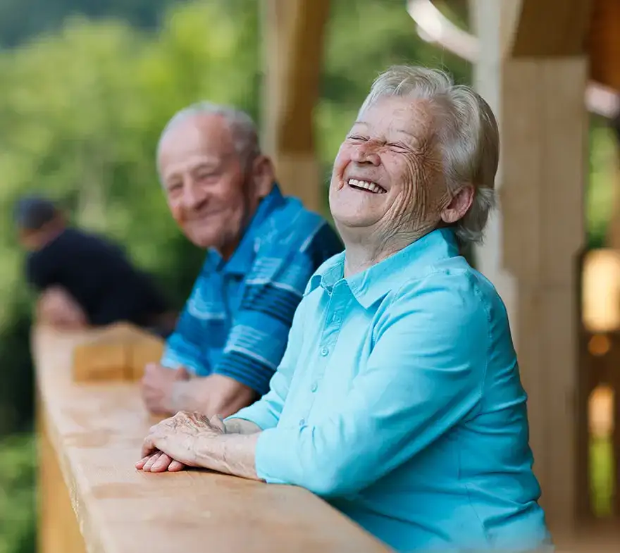Two elderly individuals smiling and enjoying time together on a wooden balcony surrounded by greenery. The woman in a light blue shirt appears joyful, while the man in a striped shirt sits nearby.