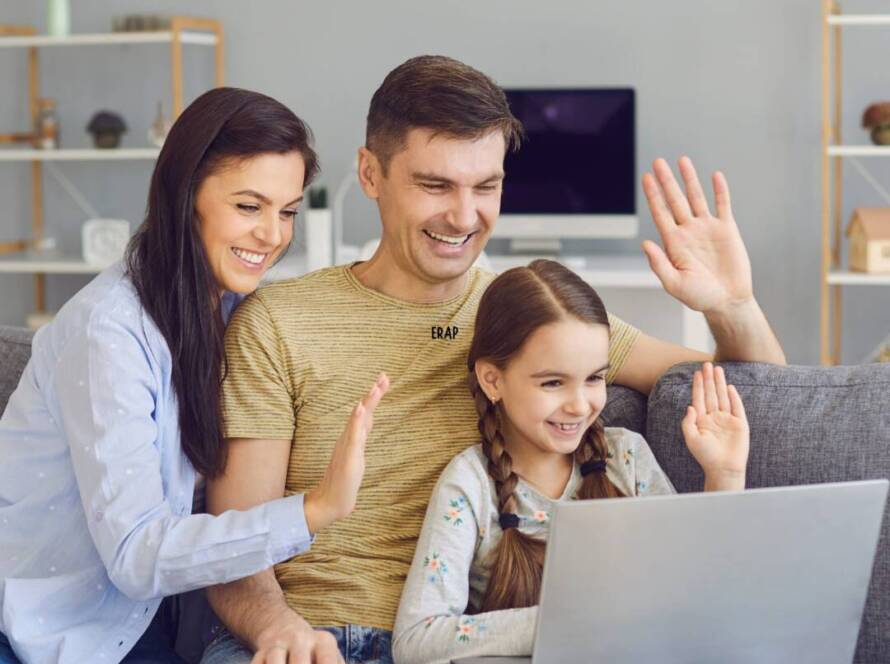 Familia de madre, padre e hija sentados en el sofá de su casa, saludando a la pantalla de una laptop