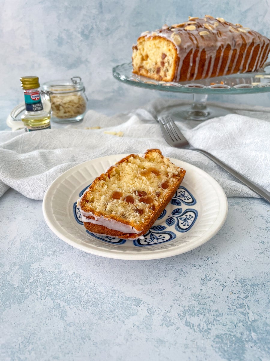 slice of cherry loaf cake on a small blue patterned plate. Rest of the cake on stand in the background.