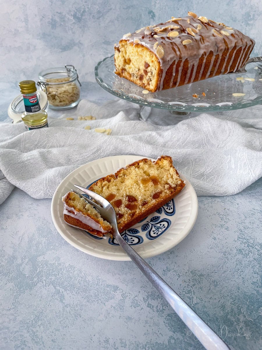 slice of cherry loaf cake on a small blue patterned plate. Rest of the cake on stand in the background.
