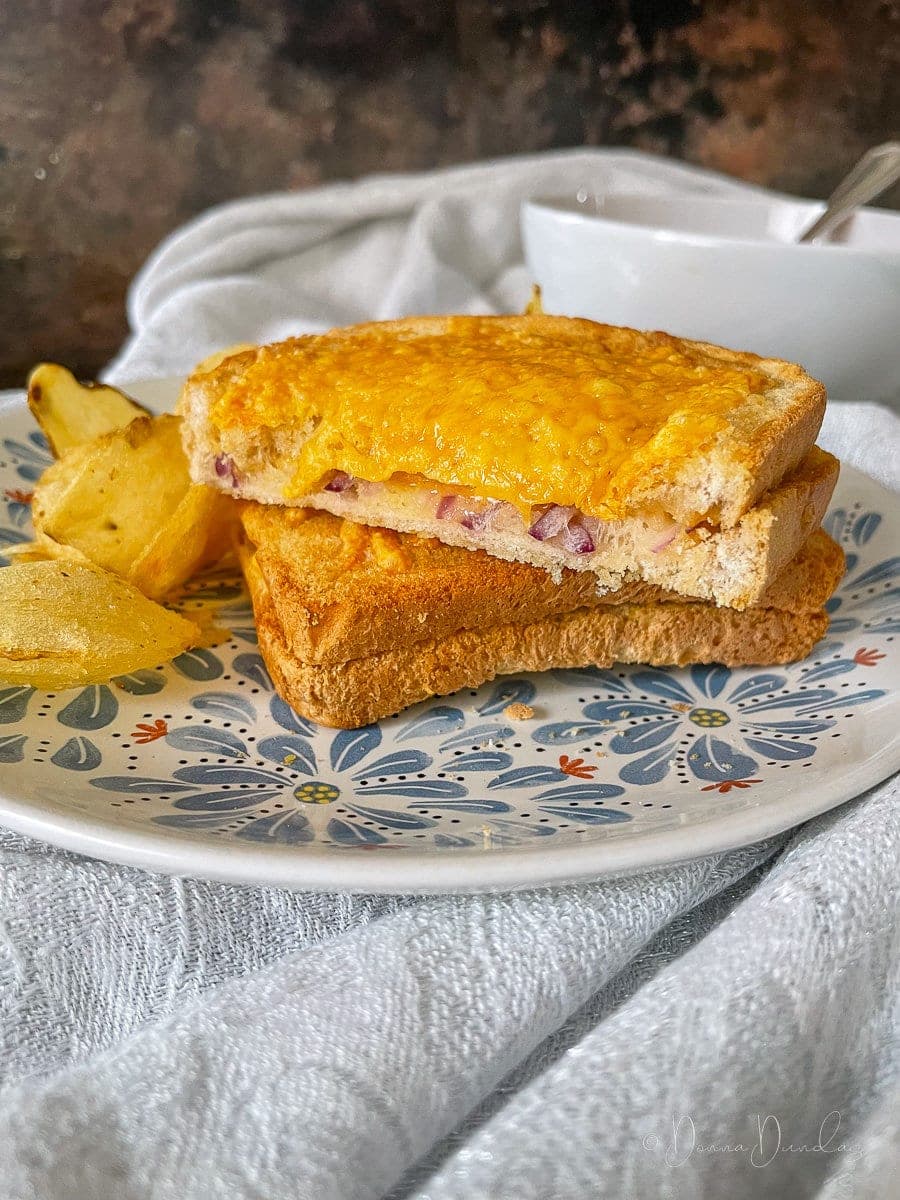 air fryer cheese an onion toastie cut in half and served on patterned plate with crispy. Bowl of soup in the background. Onions visible in the middle.