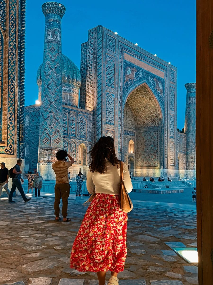 Woman with a red skirt admiring an imposing madrasah with two flanking minarets and a turquoise dome, starting to light up after sunset