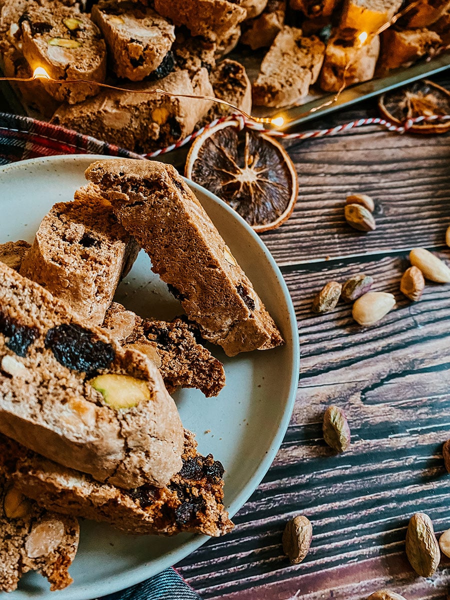 christmas biscotti on a wooden background, some on a plate and some on a tray, around fairy lights, pistachios and dried orange slices