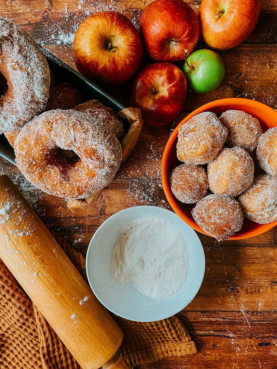 wooden background, apple cider donuts, a rolling pin, a napkin, sugar and apples on the top side