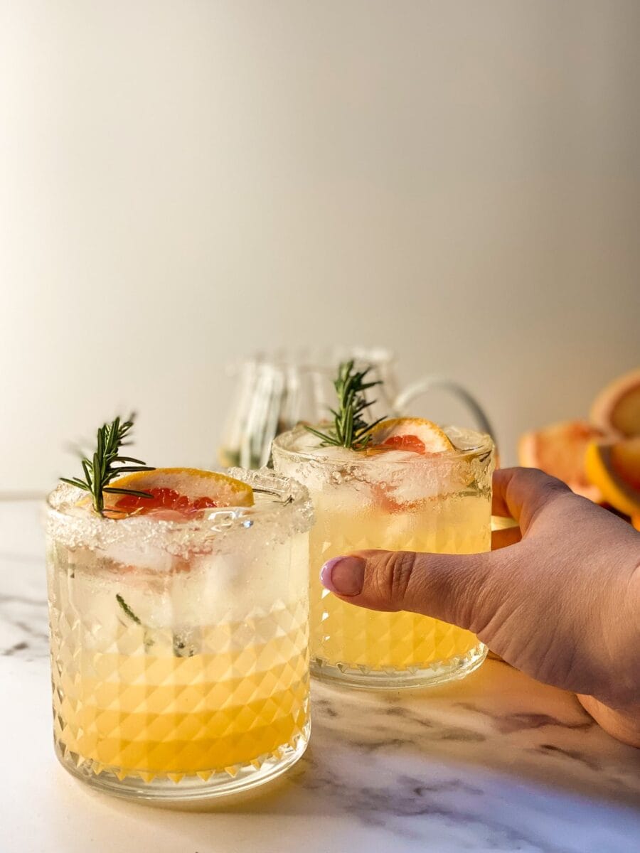 two glasses containing a grapefruit mocktail with rosemary, on the back a glass carafe and grapefruits