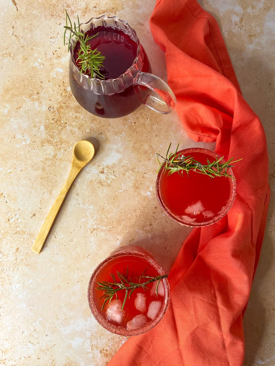 two glasses with the love potion drink with a rosemary twig as garnish, a glass jug of juice on the back, a wooden spoon on the left and a coral red napkin on the right