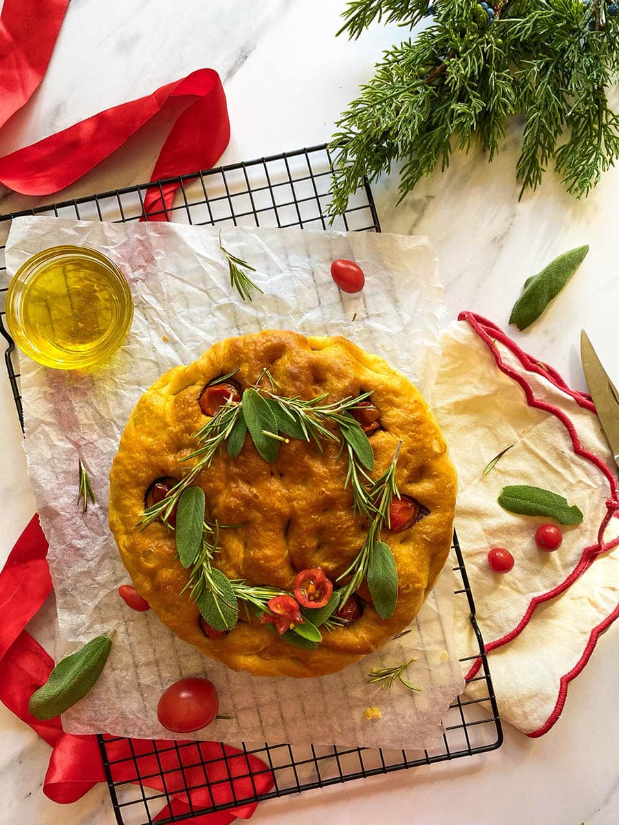 christmas focaccia on parchment paper on top of a wire rack, tomatoes, sage and rosemary around, towels, ribbons and fir on marble background