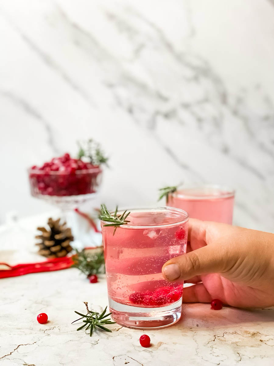 hand reaching for a glass of two glasses of mistletoe kiss, in the background cranberries, white and red towel, pinecones, around cranberries and rosemary, on marble background