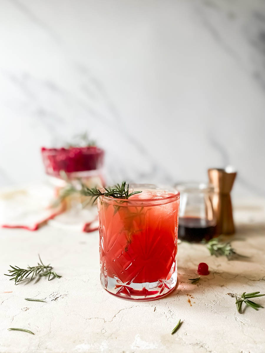 tipsy spritzer in a glass with a rosemary sprig, more rosemary and cranberries around, towel, cranberries in a pot and a jug of syrup in the back, on marble background