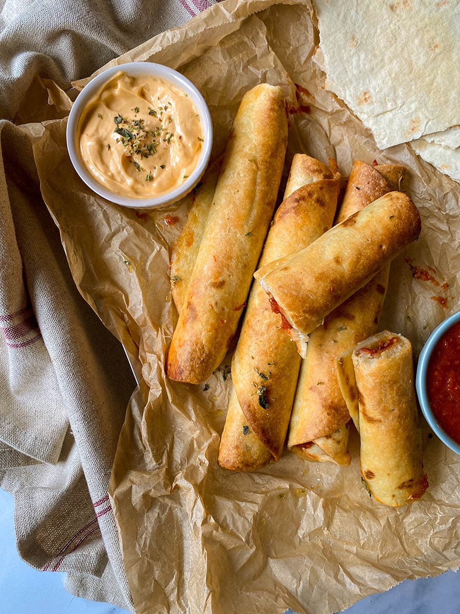 air fryer pizza roll ups on parchment paper, beside two bowls with sauces, more tortilla wraps and a towel, from above