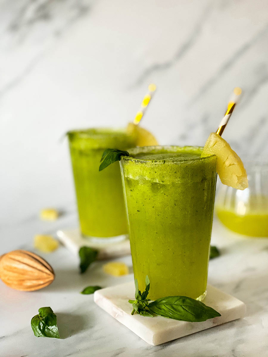 two glasses of pineapple basil mocktail on marble coasters, behind a jug with juice, around a lemon squeezer, pineapple bits and basil leaves, on marble background