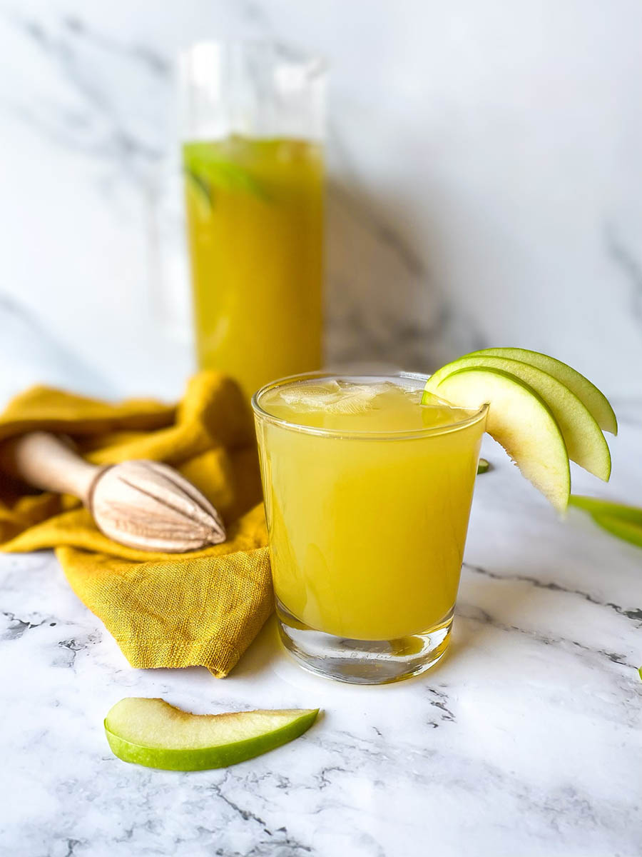 on a marble background, a glass of green apple mocktail, beside a yellow towel and lemon squeezer, behind a jug of green apple mocktail, slices of apples around