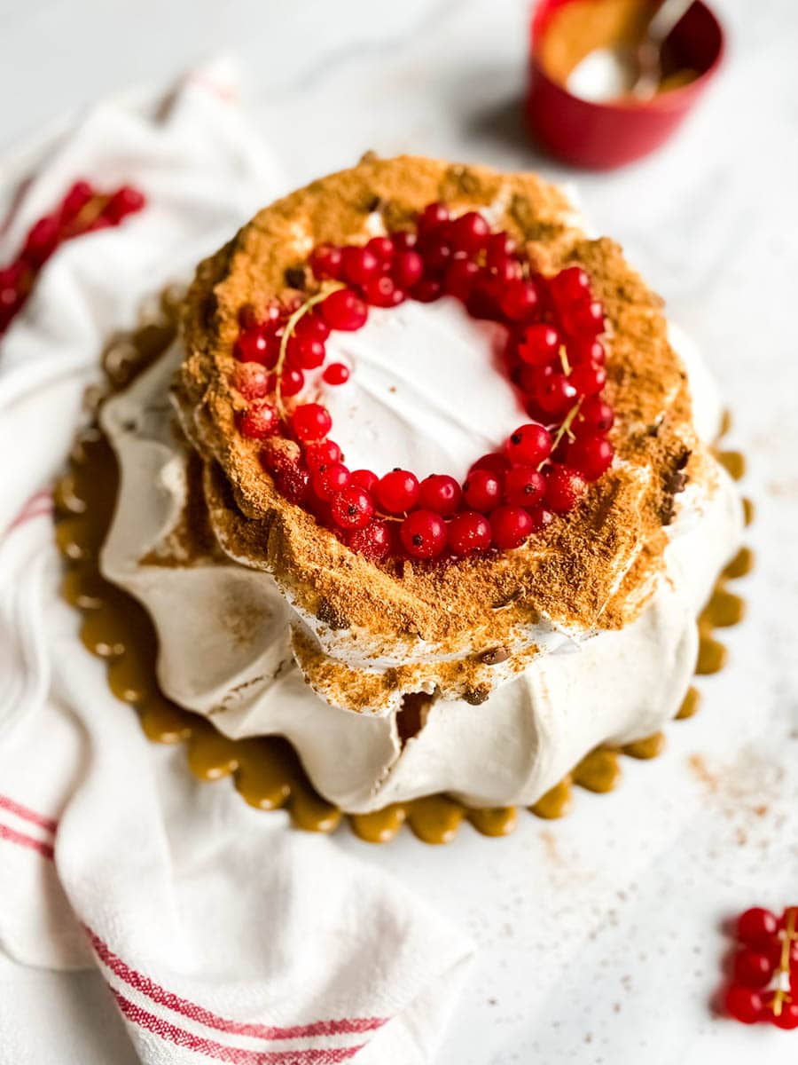 A beautifully styled gingerbread pavlova topped with a wreath of vibrant red currants sits on a white surface. The dessert features a crisp, meringue-like base with a dusting of gingerbread crumbs, creating a rustic yet elegant presentation. A red and white striped cloth adds a festive touch to the composition.