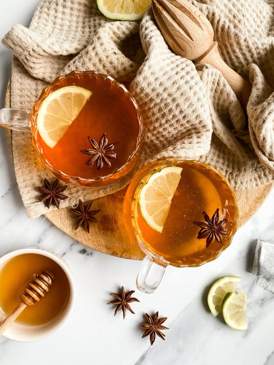 from top two cups of hot toddy mocktail with slices of lemon and star anise, over a wooden board and light brown tea towel, slices of lemons, star anise and honey around,