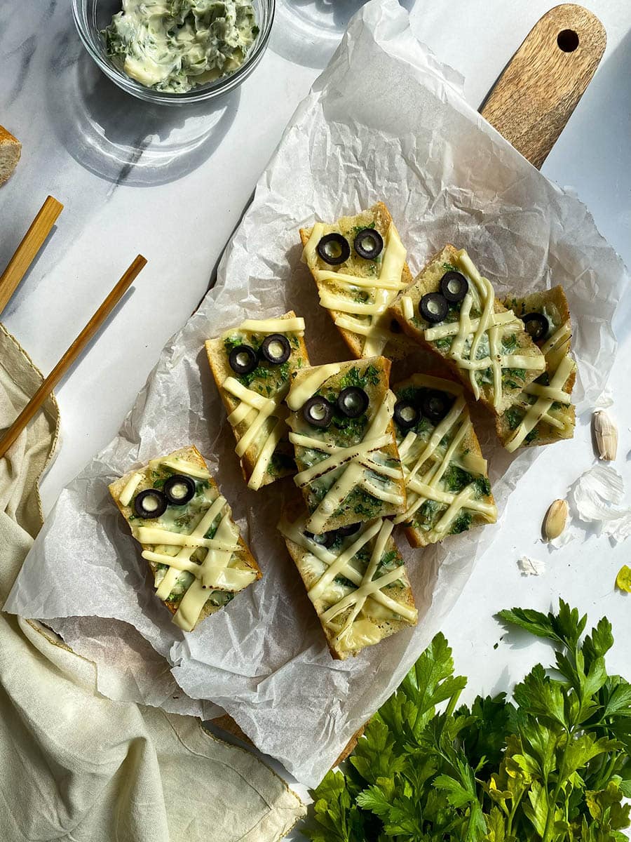 mummy garlic bread pieces on a wooden board, parsley around, a bowl with garlic butter and a bowl with olives in the back, towel and garlic around
