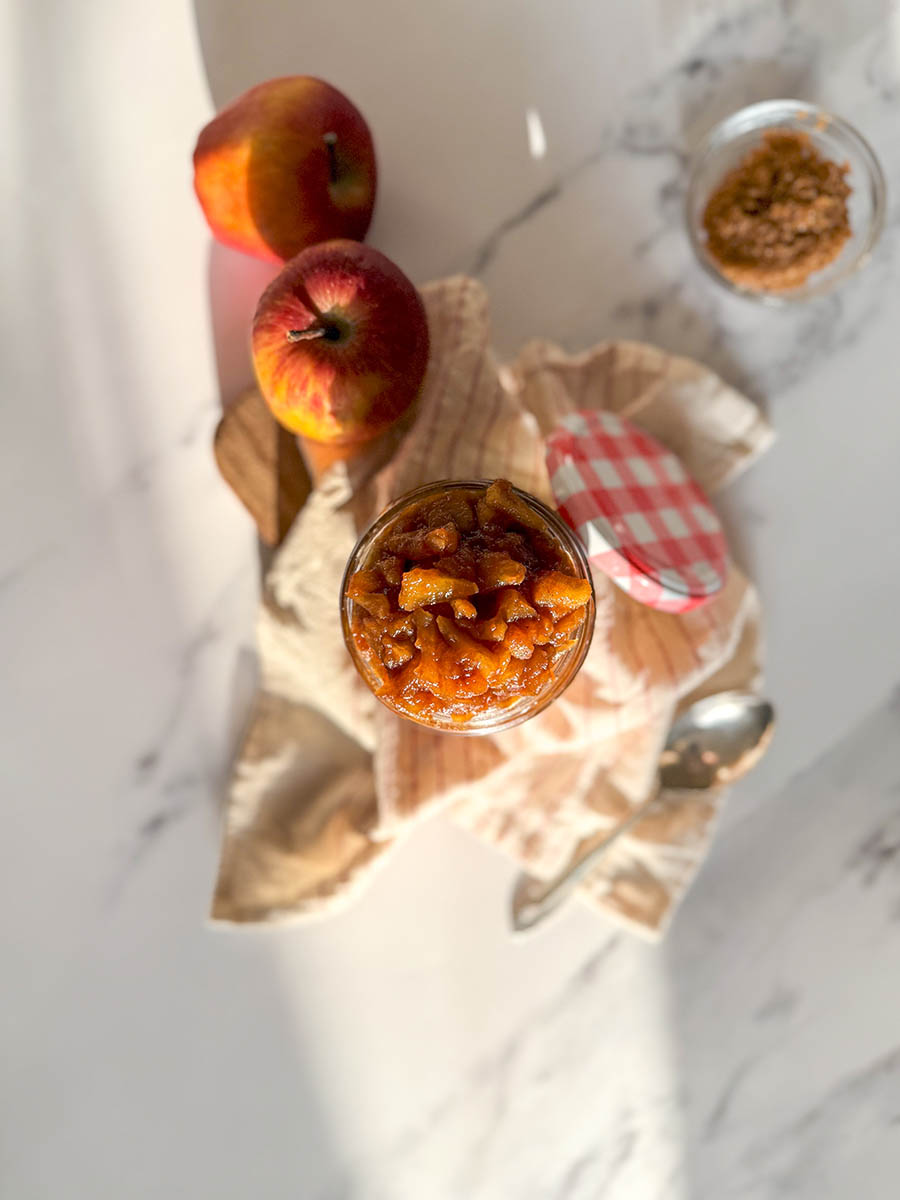 from top - apples caramelized in a jar, over a striped towel, a spoon, apples and brown sugar on the sides, over a marble background