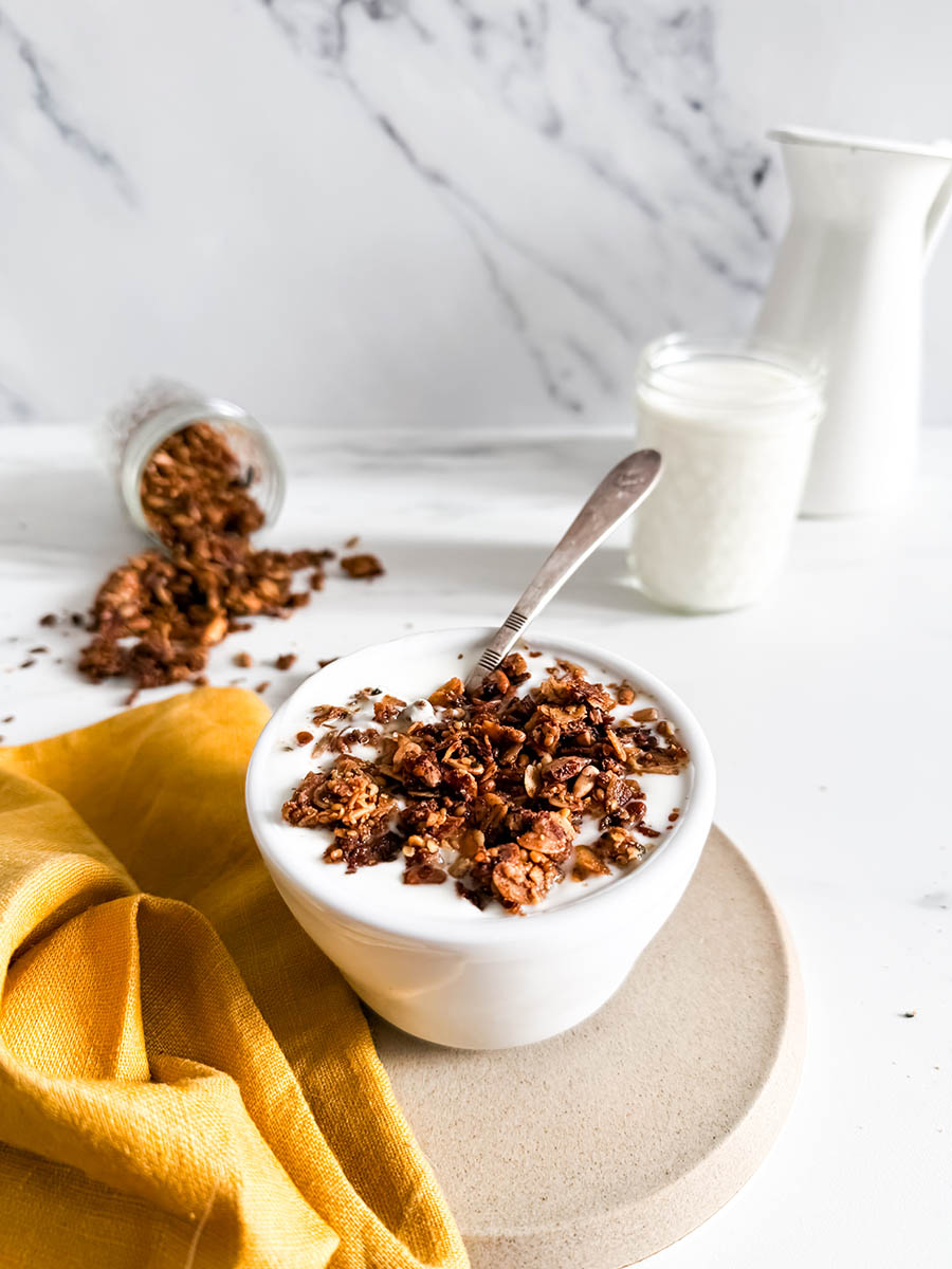 Granola with cinnamon in a bowl of creamy yogurt, placed on a simple beige plate. A vibrant yellow cloth and spilled granola enhance the cozy breakfast vibe, with a milk jar in the background.