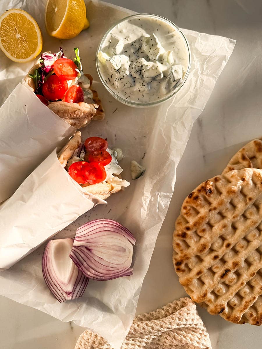 Top-down view of chicken pitas on parchment paper, filled with grilled chicken, fresh cherry tomatoes, mixed greens, and drizzled with creamy tzatziki sauce. Surrounding the wraps are a bowl of tzatziki, red onion slices, pita bread, and fresh lemon halves.