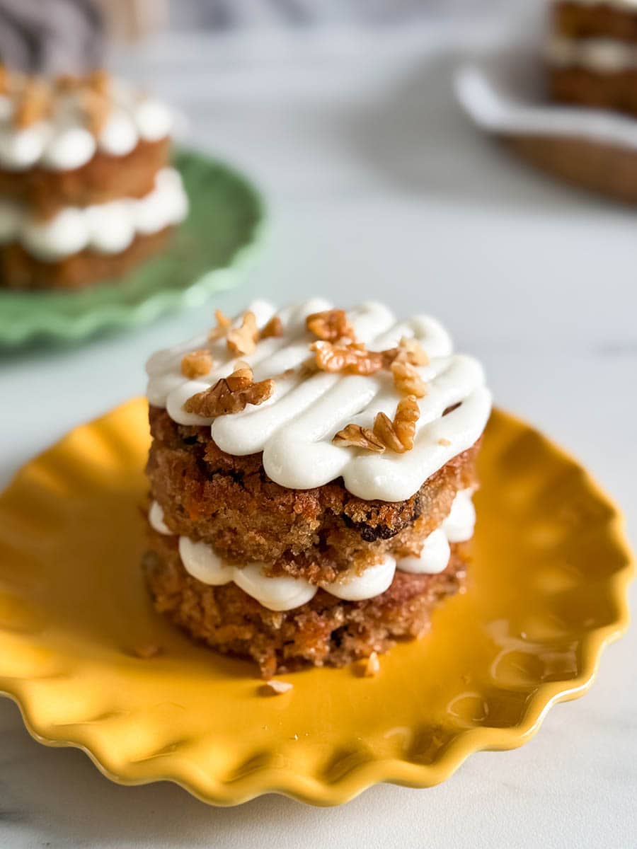 Another angle of a stacked carrot cake with creamy frosting and walnuts, placed on a yellow plate, with additional cakes blurred in the background.