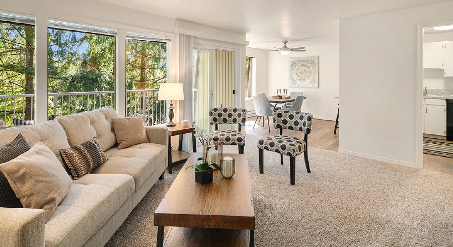 Bright living room with large sofa, patterned chairs, dining table in background, and view of trees outside windows