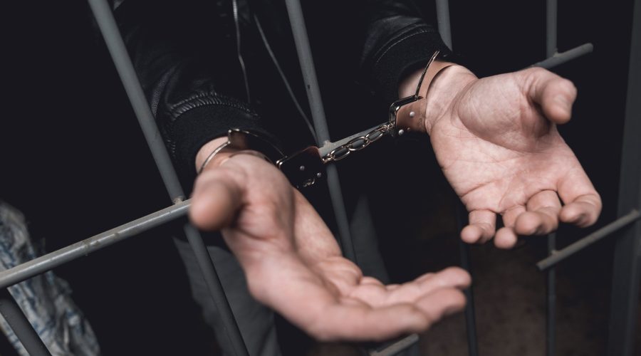 Man In Handcuffs Behind Bars In A Police Station.