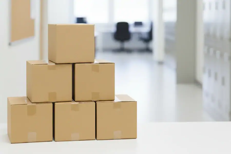 A collection of clean, stacked cardboard moving boxes sitting outside near buildings that resemble an office, a school, and a community center, illustrating common institutional places to find free boxes from heavy shipments.