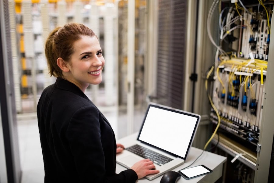 IT professional working on laptop in server room, smiling. Data center maintenance and network infrastructure.
