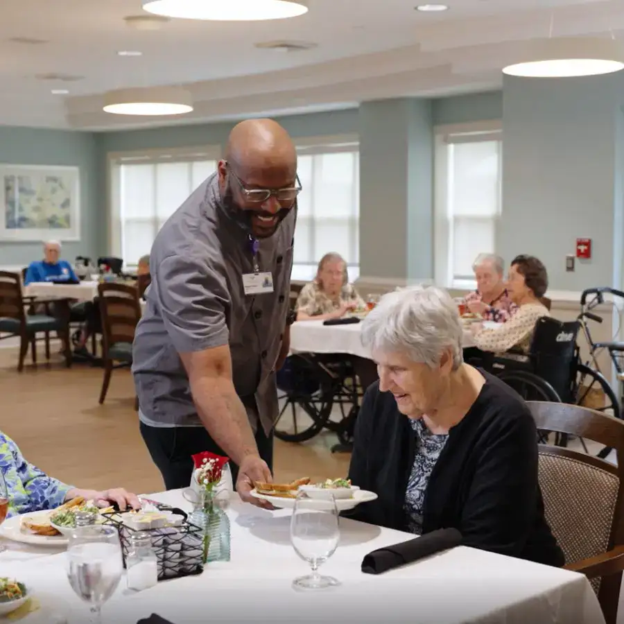 A caregiver serves a meal to an elderly woman in a dining area, with other residents enjoying their meals in the background. The setting is bright and welcoming, highlighting a community atmosphere in a senior living facility.