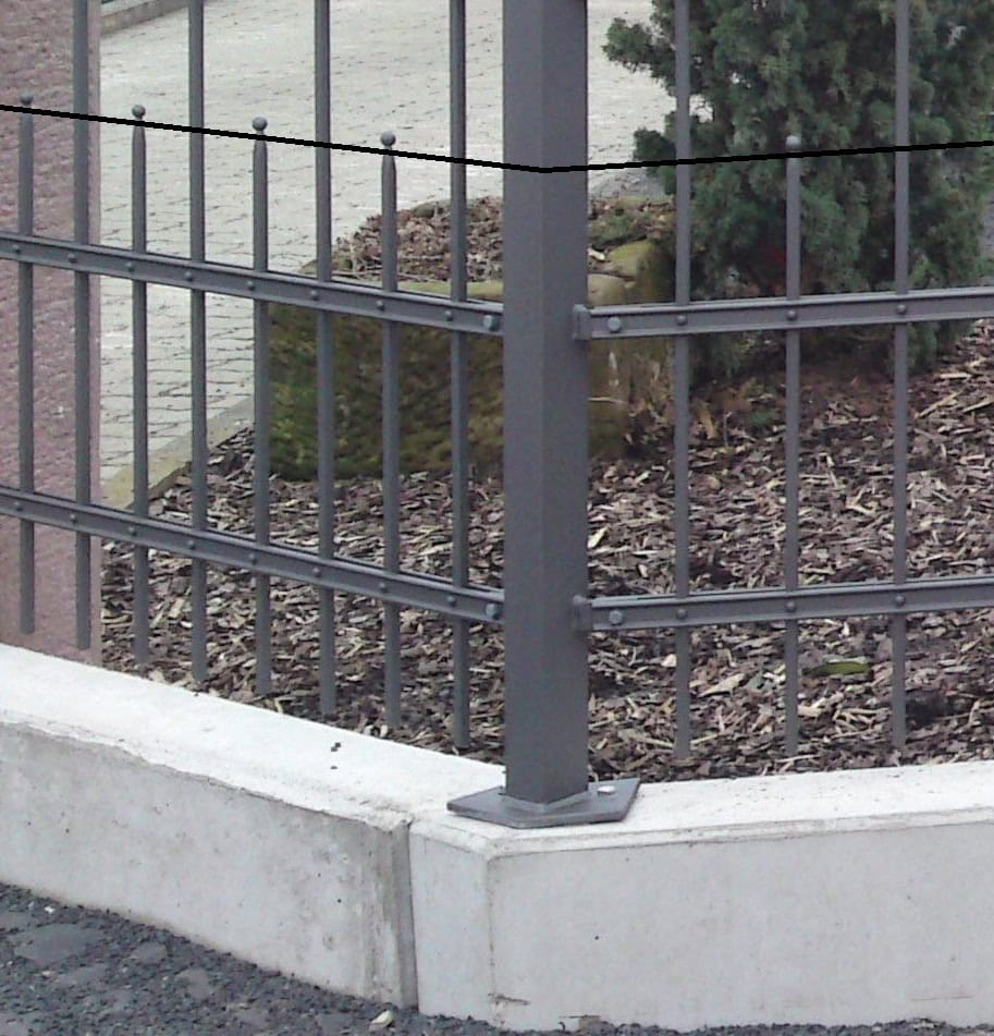 Dunkelgrauer Metallzaun von Zaunbau von Stahl- und Metallbau Drost mit vertikalen Stäben, Zierspitzen und genieteten Querstreben, auf einem Betonsockel befestigt.