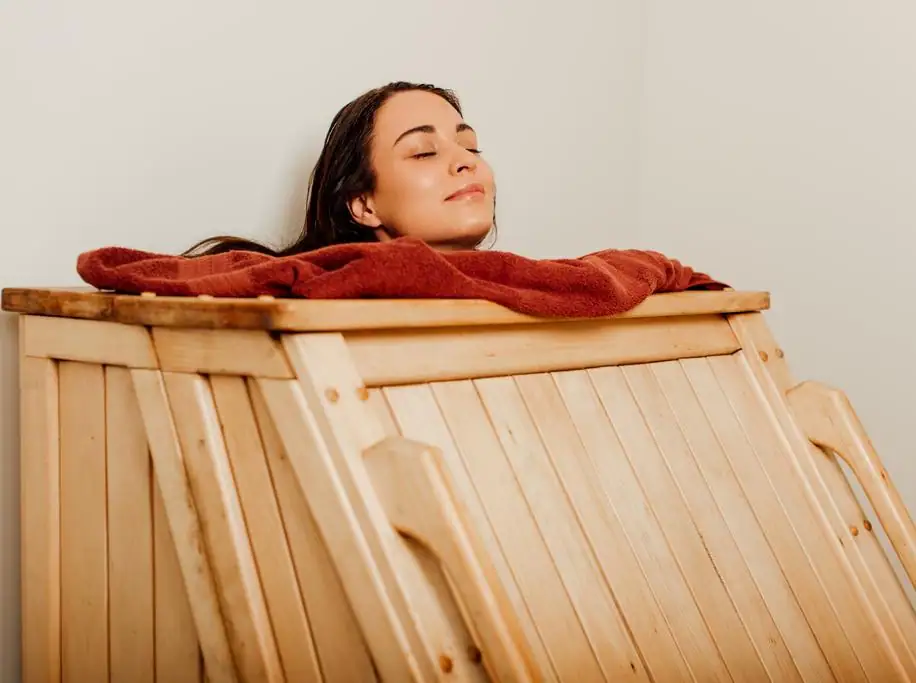 Swedhanam - An eye-level medium shot shows a woman with dark hair and a serene expression resting inside a light-colored wooden steam cabinet. Her eyes are closed and her head is tilted back slightly, resting against a plush, reddish-brown towel draped around the neck opening of the box. The cabinet is constructed from vertical wooden slats, and the warm, soft lighting creates a tranquil atmosphere against the neutral, off-white walls of the room.