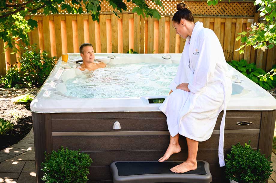 Filtre de spa: comment bien l'entretenir ? Un homme est installé dans un spa et une femme en peignoir est installé sur le rebord de l'équipement et regarde le panneau de commande.