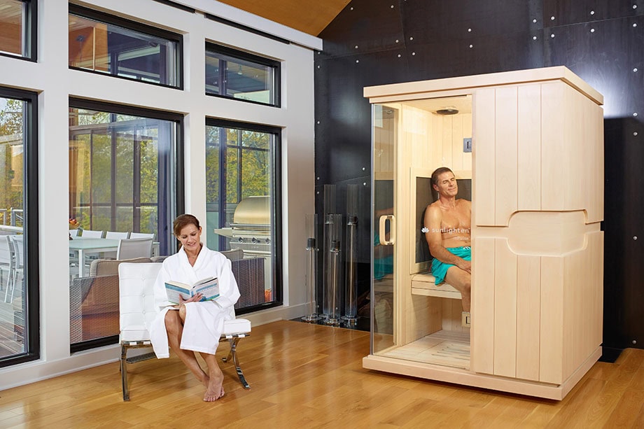 Un homme est assis sur le banc d'un sauna infrarouge installé dans une pièce à vivre. Une femme en peignoir est assise sur une chaise à côté de l'équipement et lit un livre.
