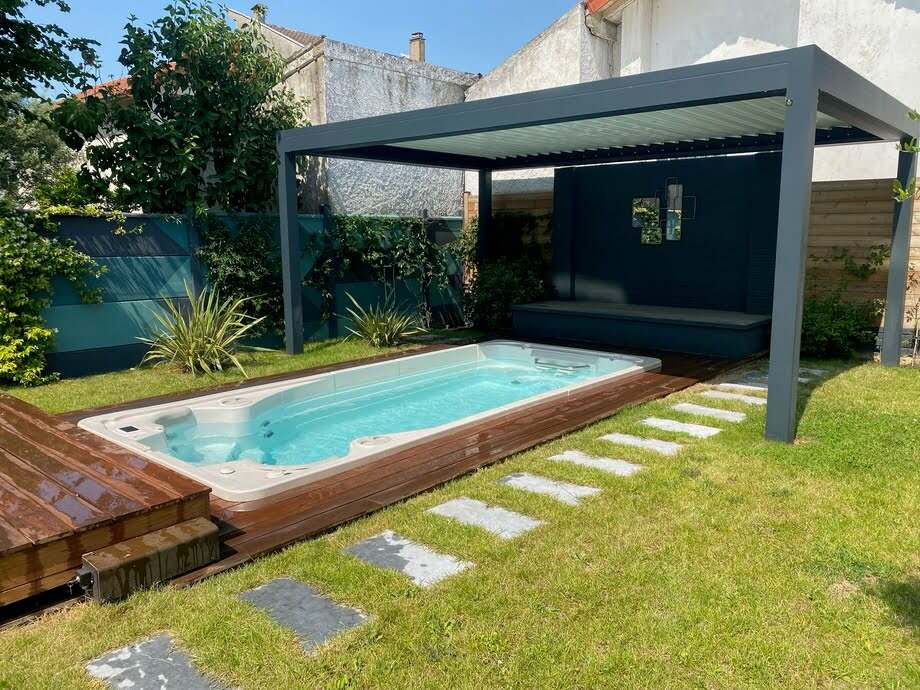 Un spa de nage est installé dans un jardin, à proximité d'une pergola bioclimatique. 