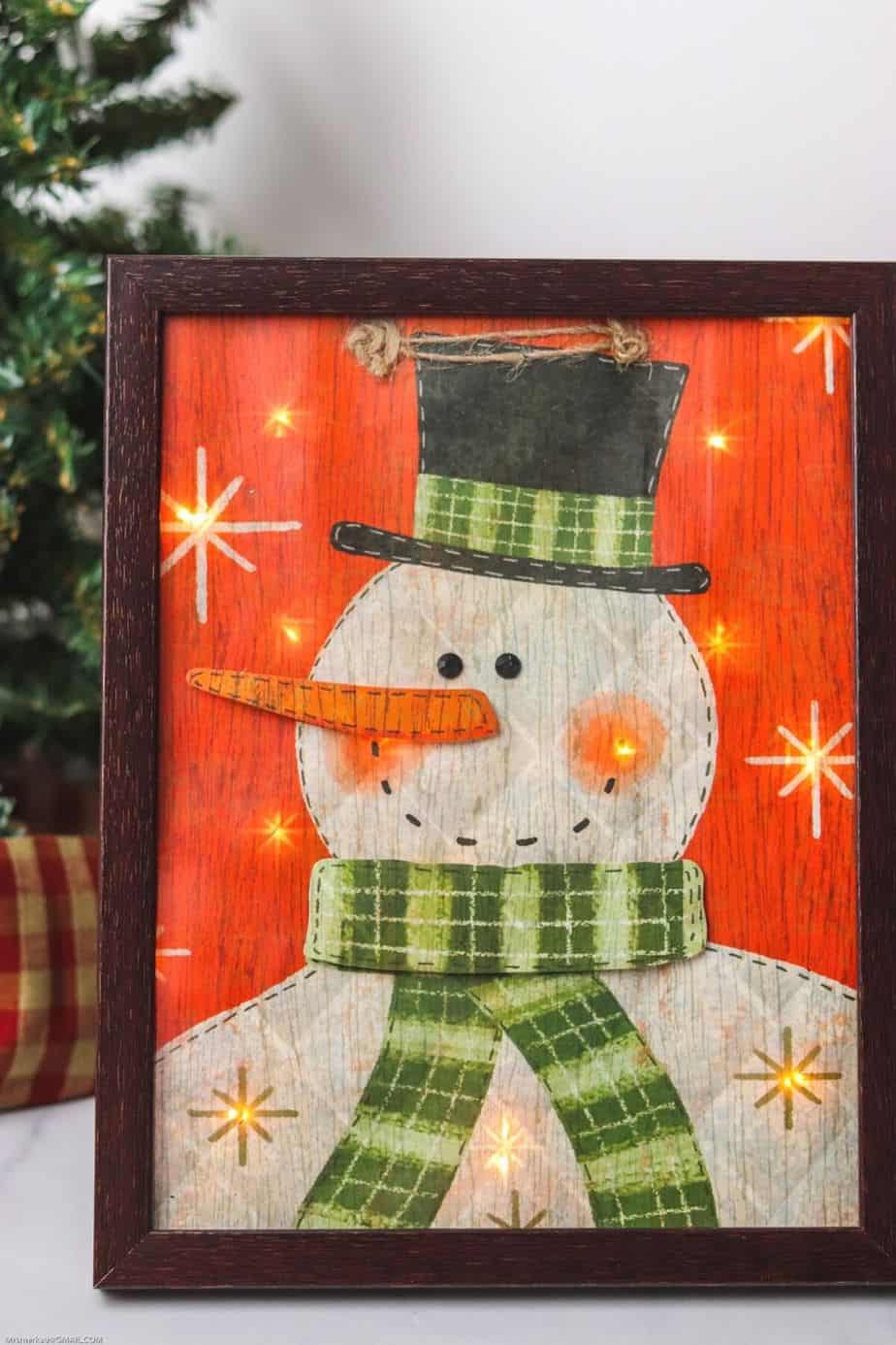 Decorate your home for the holiday season with this easy-to-make Lighted Snowman Picture. It’s fun to put together and perfect for hanging up around the home to get into the holiday spirit. This DIY Christmas craft is made with a gift bag and a dollar store frame. It is a fun dollar store Christmas craft.