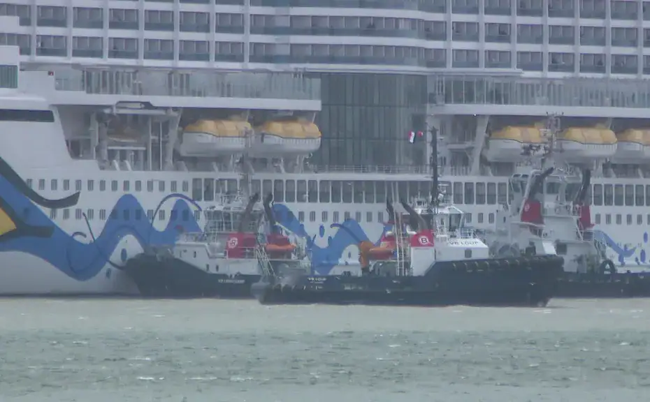 two tugboats steer beside a large cruise ship with blue wave artwork along the hull.