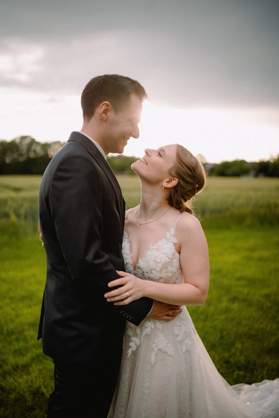 Ein Paar im Hochzeitskleid steht auf einer Wiese. Die Braut, die ein Spitzenkleid trägt, blickt zum Bräutigam im dunklen Anzug auf. Sie umarmen sich unter einem bewölkten Himmel, durch den das Sonnenlicht bricht und eine warme, romantische Atmosphäre schafft.