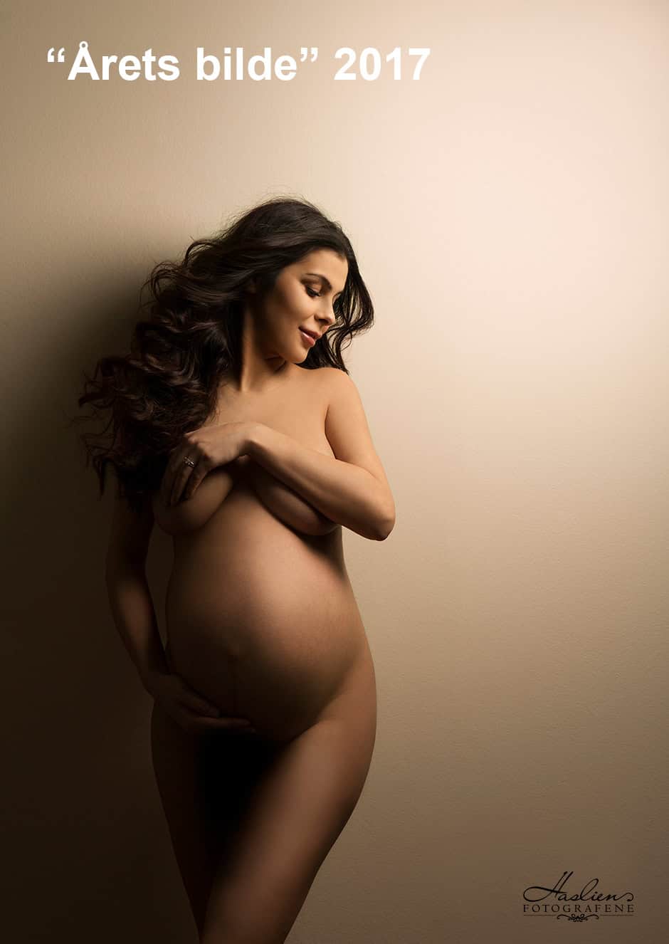 Naturlig gravidfotografi av en kvinne med lang, mørk hår som smiler og dekker brystene, vekt på gravidmage, varm og intim stemning, profesjonell gravidfoto - Gravidfotografering for minneverdige øyeblikk.