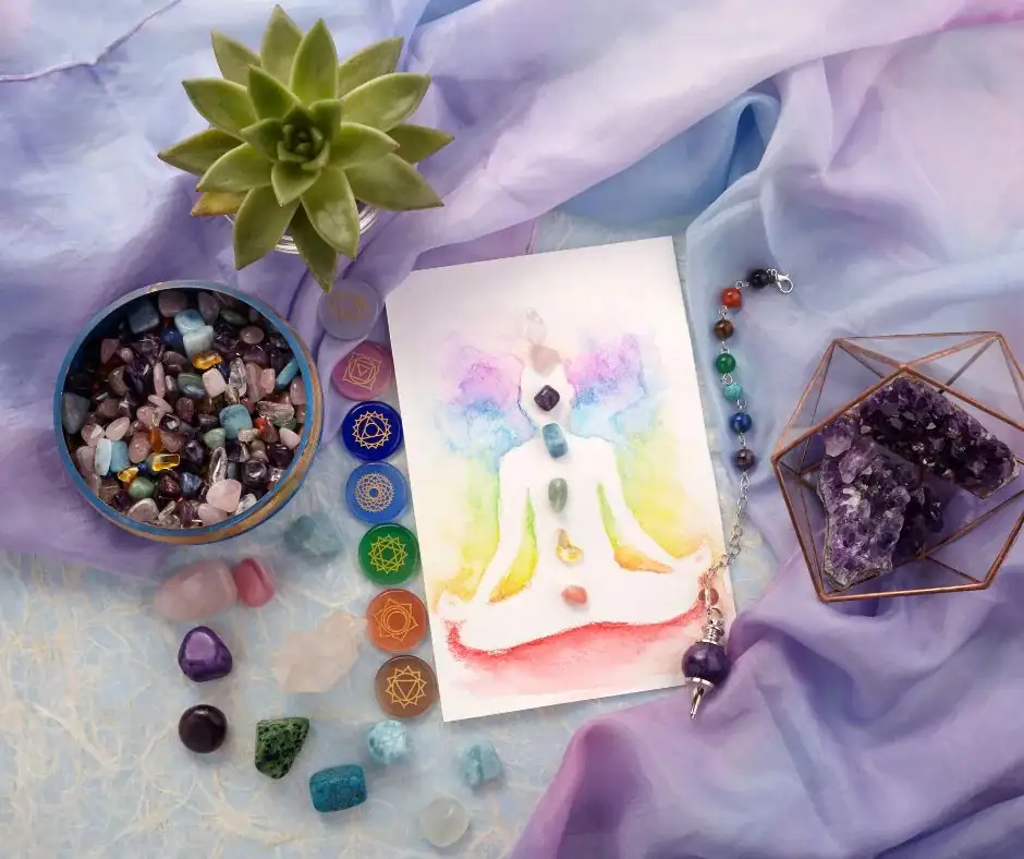 Aura Reading-A flat-lay photograph of a spiritual healing arrangement featuring a watercolor painting of a meditating figure with seven chakra stones. The scene is accented by a succulent, a bowl of tumbled crystals, an amethyst cluster in a geometric copper frame, and a chakra-symbol set, all draped in soft lavender and blue silk.