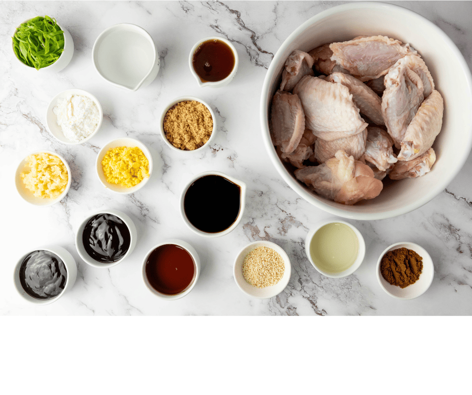 An overhead shot of raw chicken wings in a large white bowl, surrounded by various small white ingredient bowls on a white marble countertop.