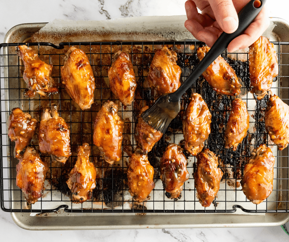 An overhead view of baked, unglazed chicken wings arranged neatly on a black wire baking rack over a foil-lined baking sheet. A hand uses a brush to baste one of the wings with sauce.