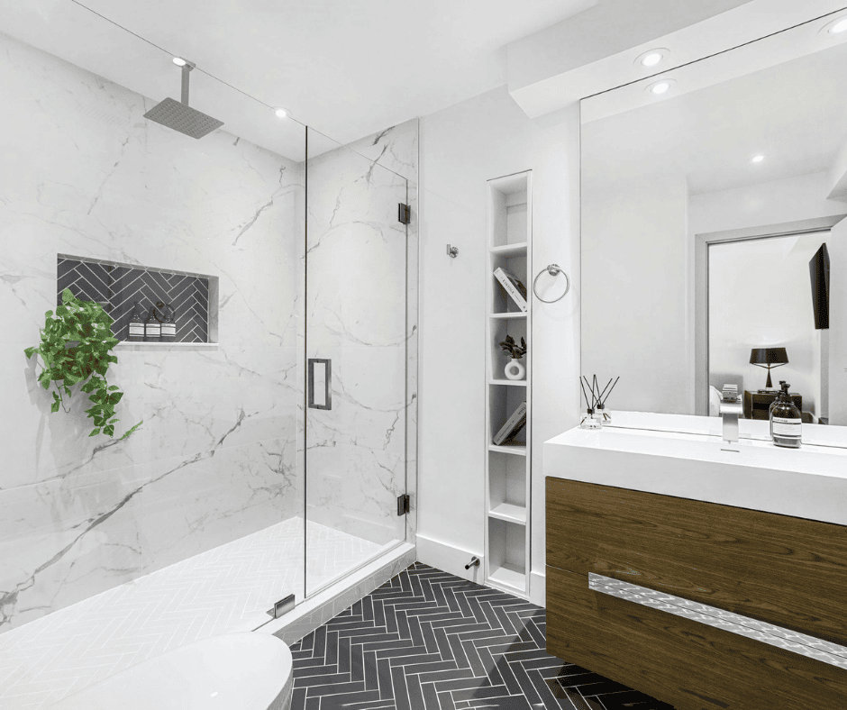 A modern bathroom featuring a walk-in shower with large white marble wall tiles and a glass door. The floor and shower base consist of dark gray subway tiles in a herringbone pattern. A floating wood vanity with a white countertop sits next to a recessed white shelving unit.