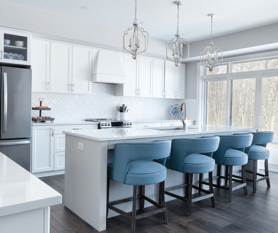 A bright, airy kitchen with white shaker-style cabinets, a white tile backsplash, and a large central island. Four cushioned light-blue barstools are lined up at the island under three decorative gold pendant lights.