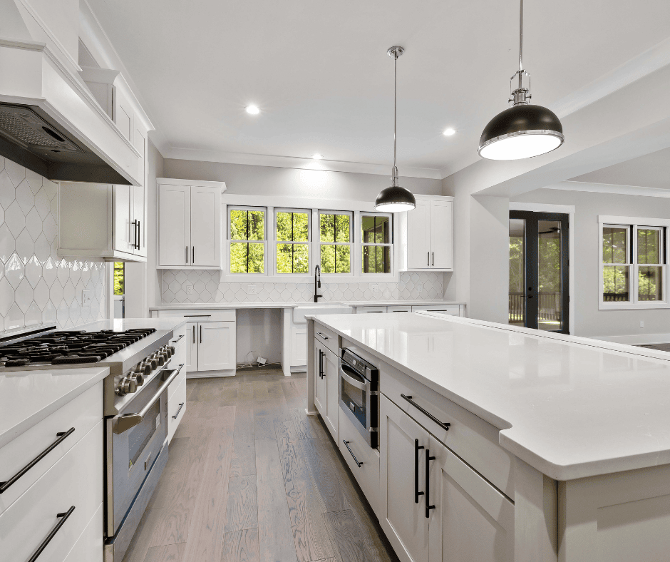 A wide-angle view of a contemporary kitchen with white cabinetry, stainless steel appliances, and expansive white quartz countertops. Two large black dome pendant lights hang over a long kitchen island, with a view of a wooded backyard through the windows.