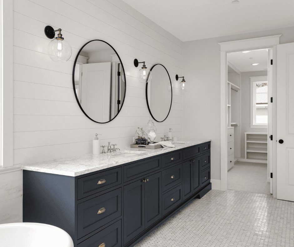 A farmhouse-style bathroom featuring a long navy blue double vanity with a marble countertop and gold hardware. Two round black-framed mirrors are mounted on a white shiplap wall, flanked by vintage-style glass globe sconces. An open doorway leads to a walk-in closet.