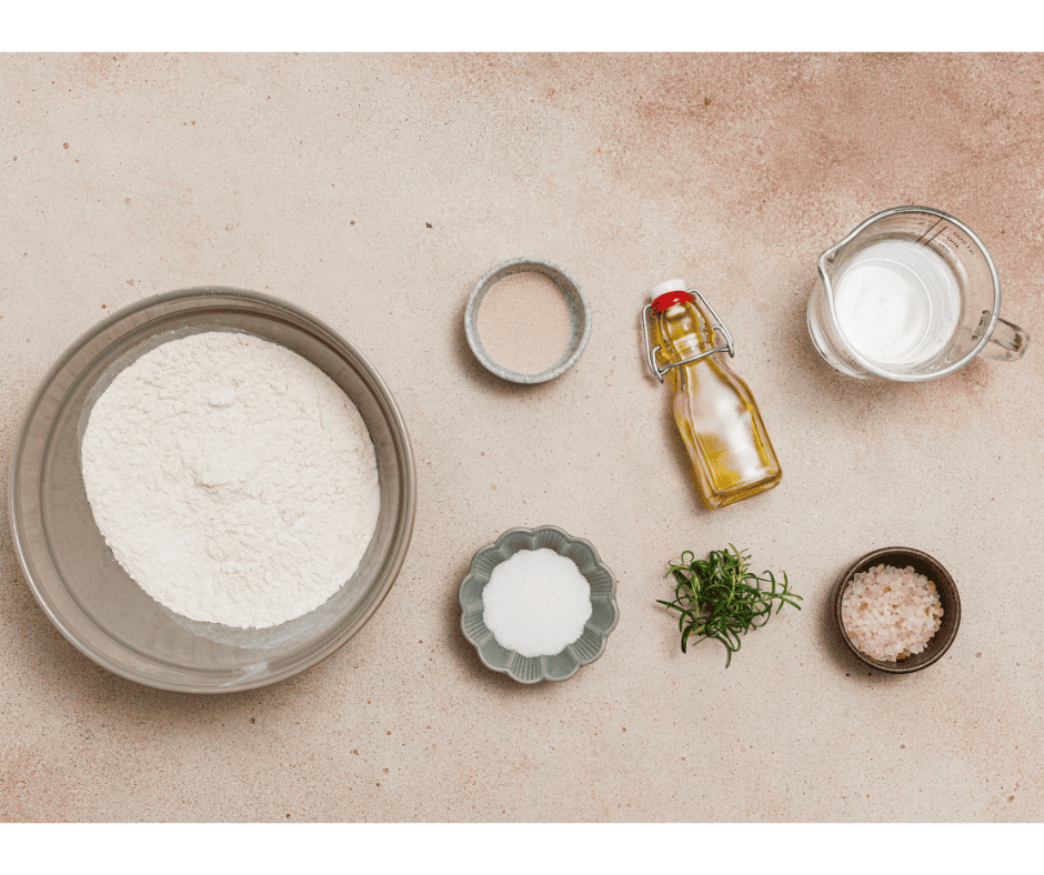 Ingredients for a focaccia bread recipe including flour, yeast, olive oil, water, sugar, and fresh rosemary on a neutral countertop.