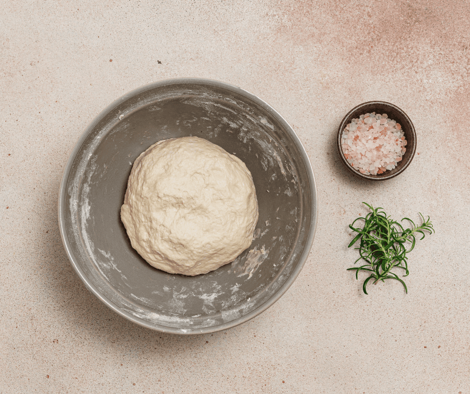 A smooth, round ball of focaccia dough in a large grey mixing bowl next to fresh rosemary.