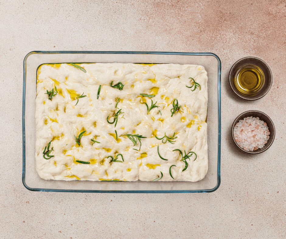 Raw focaccia dough with deep dimples, olive oil, fresh rosemary, and sea salt in a glass pan.