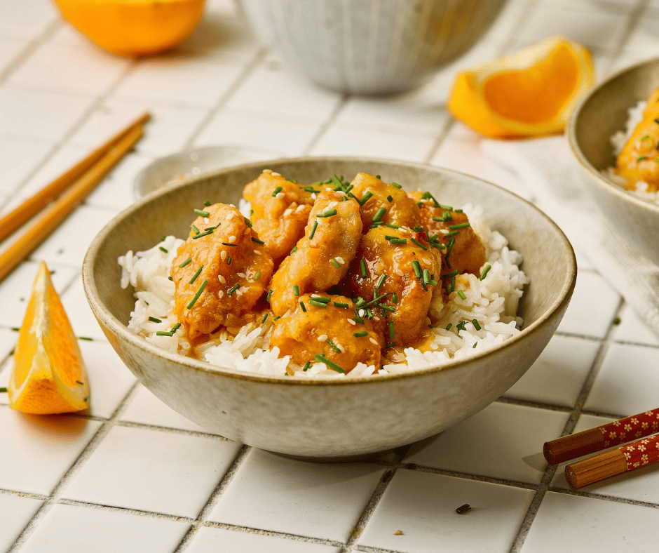 A bowl of orange chicken and white rice garnished with green onions and sesame seeds, surrounded by fresh orange wedges.