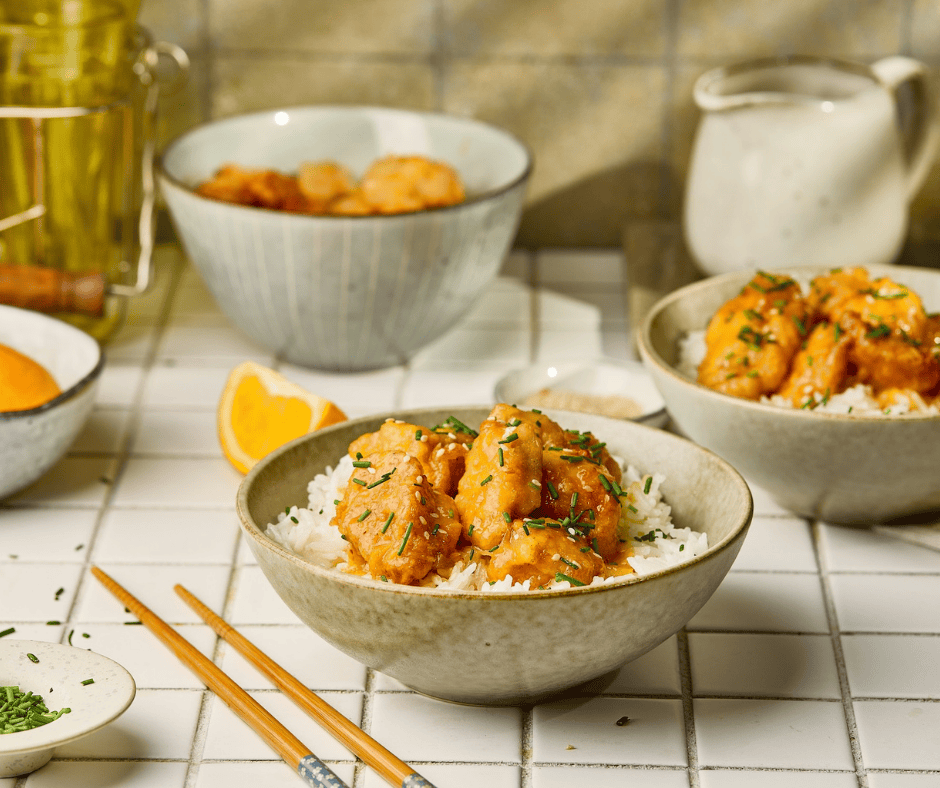 Multiple bowls of Chinese orange chicken with rice on a white tiled table with orange slices and sesame seeds.