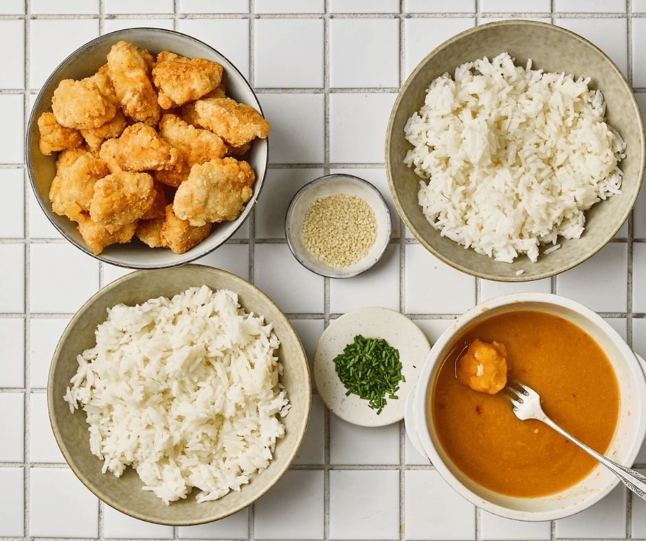 Overhead view of bowls containing fried chicken pieces, white rice, sesame seeds, chopped chives, and orange sauce.